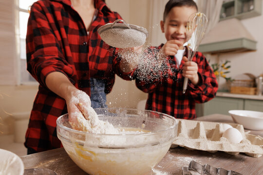Mother and son baking together in a cozy kitchen, wearing matching plaid shirts, sifting flour into a mixing bowl, joyful family cooking moment.