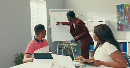 A handsome dark-skinned employee dressed in a maroon suit stands in an office next to a white...