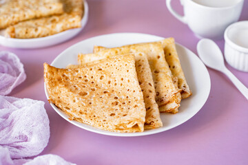 Delicious breakfast: sweet thin large pancakes on a white plate with sour cream on a pink background. Beautiful thin pancakes for dessert. Close-up