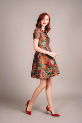 Studio portrait of beautiful redhead young woman walking in brown evening gown and red high heels....