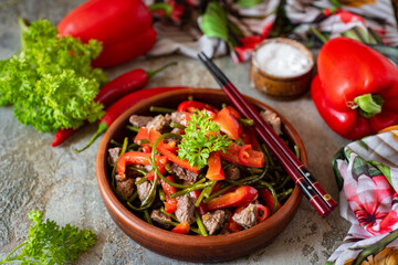 Chinese cuisine: spicy salad with meat, sweet pepper and asparagus. Close-up. Spicy salad with beef for lunch in a brown plate on a gray background. Close-up