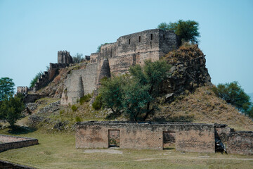 The weathered stone walls and ruins of Ramkot Fort stand atop a rugged hill, surrounded by greenery...
