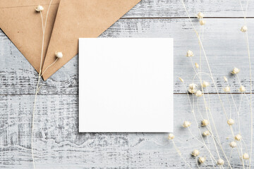 Blank square wedding invitation card mockup with dry white flax flowers decor on wooden background, top view with copy space