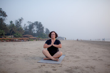 A beautiful plus size woman wearing black sports shorts and a T-shirt doing yoga on the beach