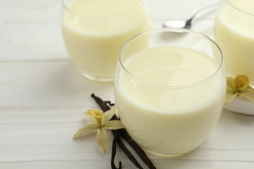 Delicious vanilla milk in glasses, pods and flowers on white wooden table, closeup