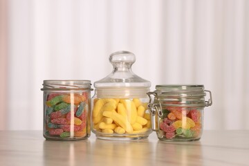 Tasty gummy candies in glass jars on white marble table