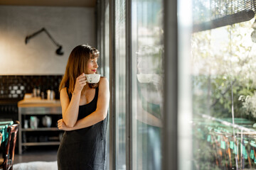 A relaxed woman in a minimalist dress leans on a rustic dining table, holding a cup of coffee. Soft...