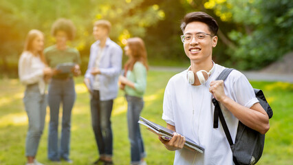 Scholarship Programs. Male asian college student with backpack and workbooks outdoors, posing at campus with classmates on background, selective focus