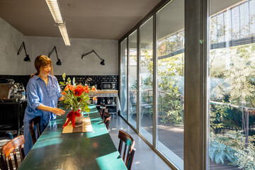 A woman in a relaxed blue shirt decorates a rustic dining table with a floral arrangement in a stylish kitchen. Natural light from large glass windows enhances the warm and inviting atmosphere