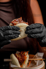 A close-up of a person wearing black gloves, holding a delicious bao bun filled with meat and...