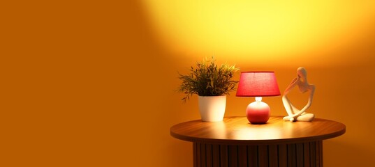 Glowing lamp and houseplant on wooden table near orange wall. Banner for design