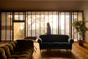 A woman in a blue outfit enjoys a peaceful morning by the large window with wooden blinds. The cozy...