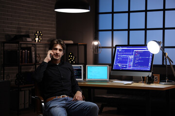 Young male programmer talking by mobile phone and working with computers in office at night