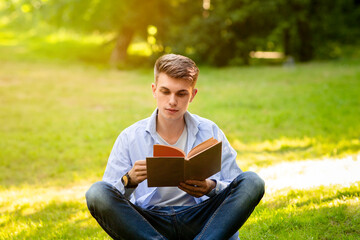 Outdoor Leisure. Young handsome guy resting with book in park, sitting on green lawn, reading new...