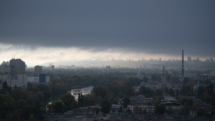 Dark clouds gather above the city skyline, casting shadows over buildings and trees as the day transitions into evening. A river reflects the changing light, creating a moody atmosphere