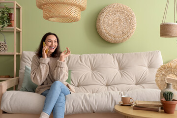 Pretty young woman talking by phone while sitting on cozy white sofa in living room