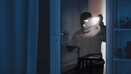 Criminal Act Concept. Portrait of robber checking apartment through balcony door, using flashlight...