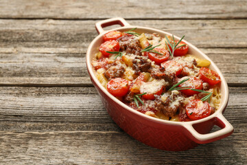 Delicious pasta casserole with cheese, tomatoes, minced meat and rosemary in baking dish on wooden table, closeup. Space for text