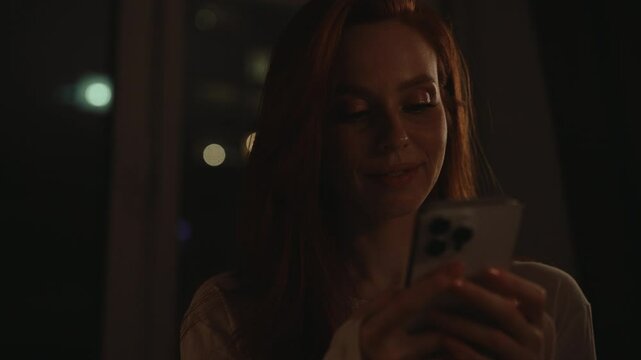 Happy young woman with long red hair smiling using mobile phone at night, sitting comfortably by window in dark living room, happily typing message and connecting online. Shooting in slow motion.