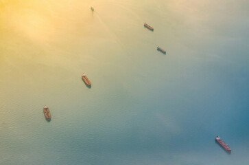 Cluster of cargo ships gently drift on calm sea waters. Warm gradient light casts serene tones, creating tranquil atmosphere. Viewed from above, scene captures peaceful maritime moment