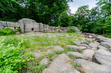 Stone dolmen located within dense forest, surrounded by fresh greenery and under overcast sky. Grey...