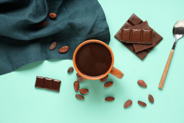 Cup of tasty hot chocolate with spoon, cocoa beans and napkin on turquoise background