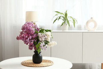 Vase with lilacs branches on table near chest of drawers in light room