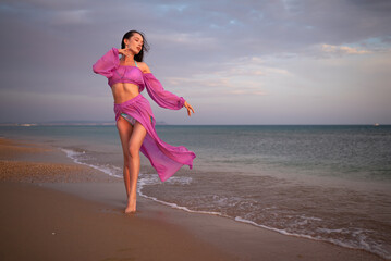 Graceful woman dancing on the beach at sunset in a flowing purple outfit