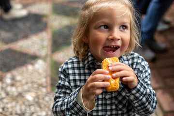 A joyful child with blonde hair and a plaid shirt, smiling with excitement while holding a snack, enjoying a bite in a beautiful outdoor setting