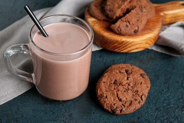 Glass cup of sweet chocolate milk and wooden board with tasty cookies on dark background