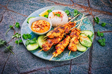 Sate ayam- grilled chicken satay sate skewers with peanut dipping spicy sauce, steamed rice and fresh cucumber served as close-up on a design plate