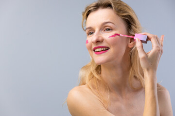 A Beautiful Woman Applying Pink Blush on Her Cheek, Showcasing Healthy Skin and Radiant Smile Against a Soft Gray Background in a Beautifying Moment of Self-Care.