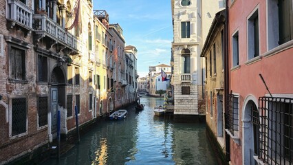 A picturesque Venetian canal flanked by historic buildings with weathered facades and elegant balconies. Small boats float on the calm water, while a distant bridge and flags add to the charming scene