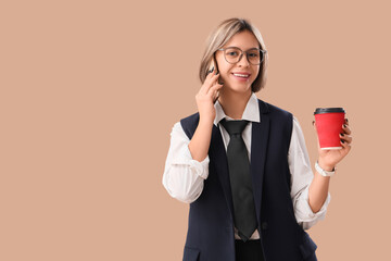 Successful businesswoman with cup of coffee talking on mobile phone on beige background