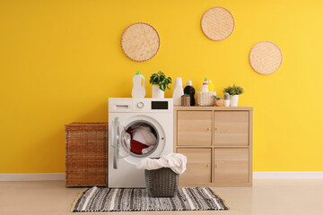 Modern washing machine, chest of drawers and laundry baskets near yellow wall in room