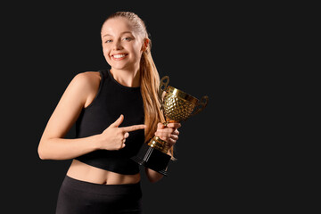 Sporty young woman pointing to golden victory cup on black background