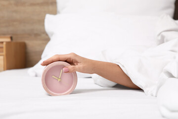 Female hand with pink alarm clock on bed in bedroom