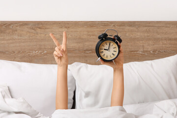 Female hands with alarm clock and showing Victory gesture in bed at home