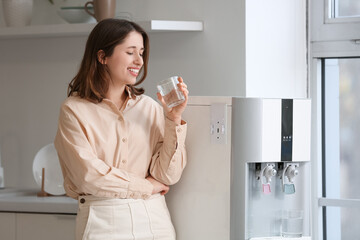 Smiling young woman drinking water from cooler in kitchen