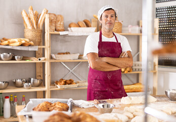 Caucasian man cook-baker stands at table in kitchen of bakery. Wide range of ready-made bread products in rack in background, rolls and croissants