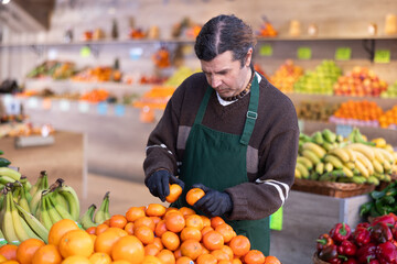 Adult man seller in apron puts fresh tangerines on display in vegetable shop