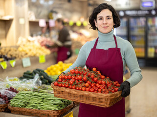 Adult woman seller in apron with basket of fresh cherry tomatoes in vegetable shop