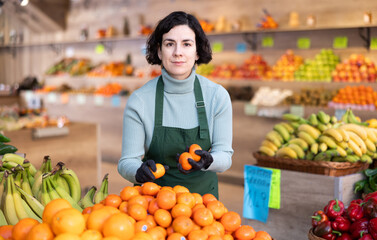 Shop seller puts goods on display case. Woman creates pyramid of tangerine, presents fresh goods. Vegetable shop near house. Spacious store with long rows of display cases, customers waiting