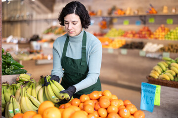 Adult female seller in uniform lays out bananas on counter in grocery store