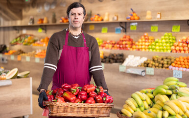 Adult man seller in apron with basket of fresh bell peppers in vegetable shop