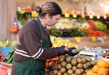 Man shop assistant puts ripe pear fruits in box on display case, arranges assortment in vegetable shop. Worker puts fruits in pile, in pyramid. Employee makes attractive display case