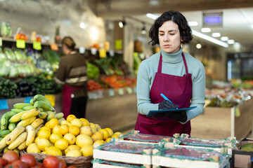 Adult female salesperson taking inventory with list in vegetable store