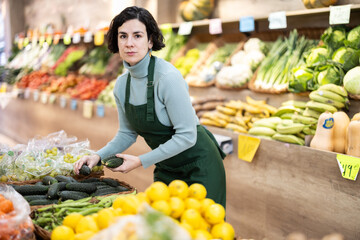 Adult woman seller in apron puts fresh cucumbers on display in vegetable shop