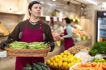 Man in apron carries large quantity of beans in pods in wicker basket to sales area, supplementing stores assortment with fresh vegetables