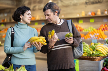 Couple adult man and woman buyers chooses bunch of fresh bananas in vegetable shop